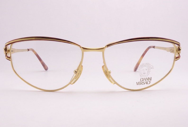 May include: Gold-tone metal eyeglasses with a brown accent on the top of the frame. The glasses have a cat-eye shape and the brand name "GIANNI VERSACE" is printed on the right lens.