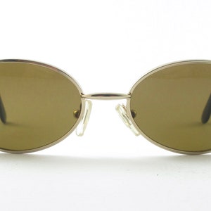 Puede incluir: Un par de gafas de sol ovaladas con montura de metal plateado y lentes marrones. La montura tiene una fina franja roja en la parte superior e inferior.