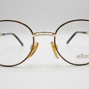 May include: Gold-tone metal framed glasses with brown temples and clear lenses. The brand name "HILTON" is printed on the right lens.