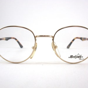 Puede incluir: Gafas ovaladas con montura dorada, puente de nariz de carey y la palabra "STING" en un diseño de estrella en la lente derecha.
