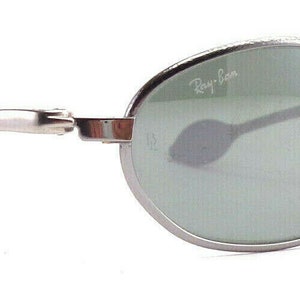 May include: Silver-framed sunglasses with oval lenses and a subtle Ray-Ban logo. The lenses are a muted green, and the temples have a sleek, curved design with dark gray ear pieces. A classic eyewear accessory.