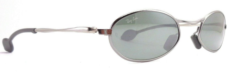 May include: Silver-framed sunglasses with oval lenses and a subtle Ray-Ban logo. The lenses are a muted green, and the temples have a sleek, curved design with dark gray ear pieces. A classic eyewear accessory.