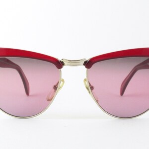 Könnte beinhalten: Rote Cat-Eye-Sonnenbrille mit goldenem Metallrahmen und rosafarbenen Gläsern. Die Sonnenbrille hat den Schriftzug "TOMMY" auf dem rechten Bügel.