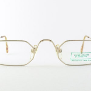 Puede incluir: Gafas de color plateado con montura rectangular y lentes transparentes. Las gafas tienen una etiqueta que dice "United Colors of Benetton. Lentes de demostración solamente."