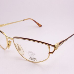 May include: Gold-tone metal eyeglasses with a brown tortoiseshell temple and a gold Medusa head logo on the side. The glasses have a clear lens.