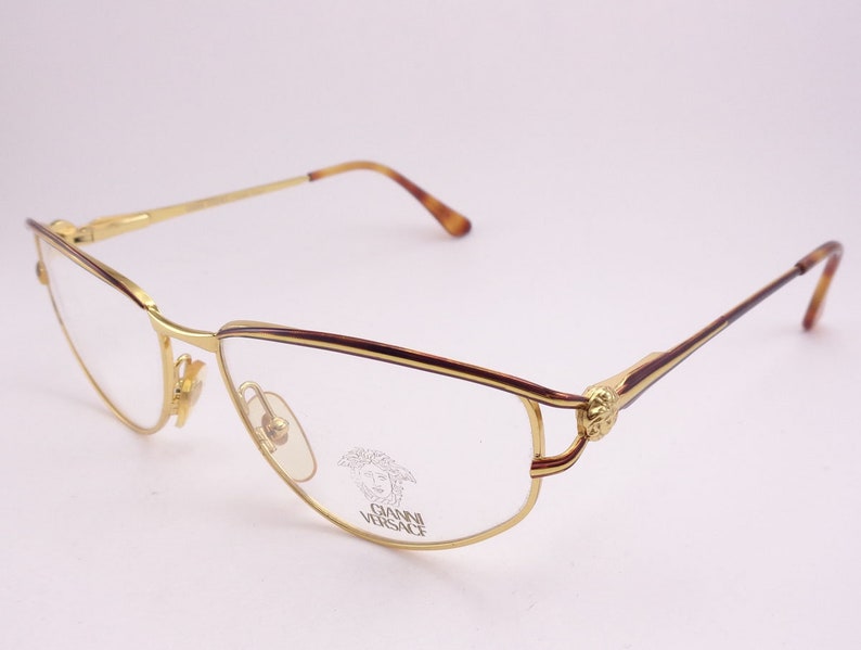 May include: Gold-tone metal eyeglasses with a brown tortoiseshell temple and a gold Medusa head logo on the side. The glasses have a clear lens.
