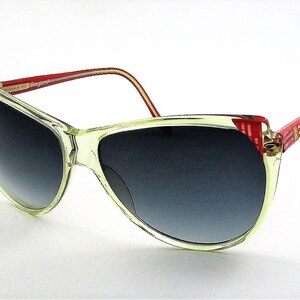 Puede incluir: Un par de gafas de sol transparentes y claras con detalles rojos y blancos en las patillas. Las lentes son de color gris oscuro.