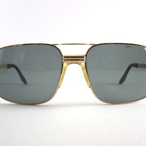 Może przedstawiać: Okulary przeciwsłoneczne typu aviator w kolorze złotym z szarymi szkłami. Okulary mają napis "Safilo" nadrukowany na lewej szklance i "Safilo" nadrukowany na prawej szklance.