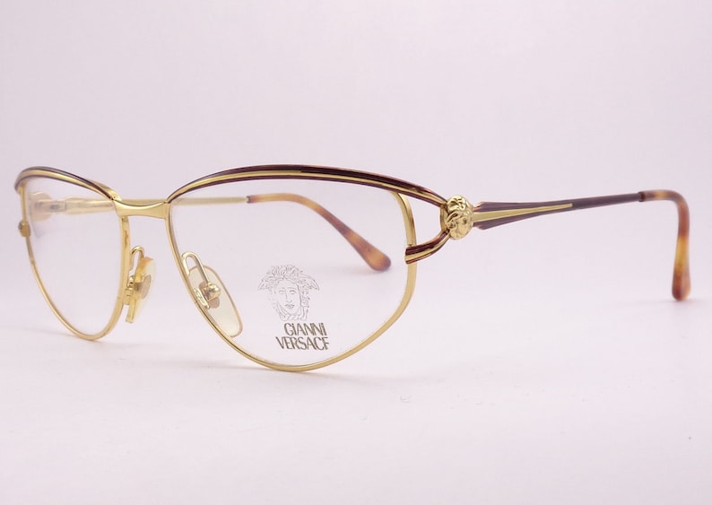 May include: Gold-tone metal eyeglasses with a tortoiseshell frame and a small, engraved logo that says "Gianni Versace" on the right lens.