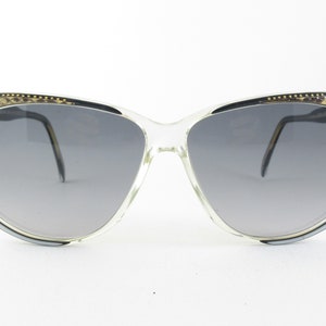 Puede incluir: Gafas de sol de plástico transparente con un patrón de piel de serpiente marrón y negro en la parte superior del marco. Las lentes son grises.