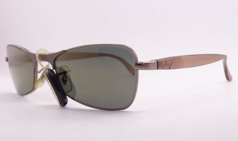 Puede incluir: Un par de gafas de sol con montura de metal marr&oacute;n y lentes verdes. Las gafas tienen el logotipo de Ray-Ban en la patilla derecha.