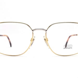 Pode incluir: Óculos de metal dourado com ponte prateada e lentes transparentes. O nome da marca "GD Gerald Genta" está impresso na lente direita.