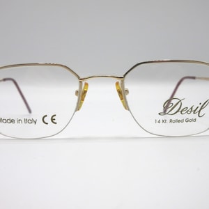 Op de afbeelding: Gouden metalen bril met een rechthoekig montuur. De bril heeft een helder glas en de woorden "Desil 14 Kt. Rolled Gold" zijn gedrukt op het rechter glas. De woorden "Made in Italy CE" zijn gedrukt op het linker glas.