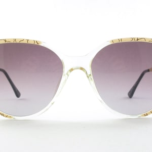 May include: Clear plastic sunglasses with a gold patterned frame and gradient gray lenses.