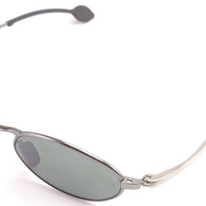 May include: Oval-shaped sunglasses with a silver metal frame and gray lenses. The temples are silver with gray rubberized ear pieces. The sunglasses are shown against a white background.