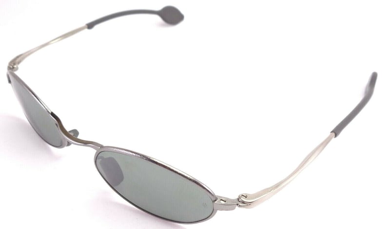 May include: Oval-shaped sunglasses with a silver metal frame and gray lenses. The temples are silver with gray rubberized ear pieces. The sunglasses are shown against a white background.