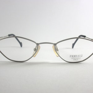 Może przedstawiać: Srebrne okulary metalowe w kształcie kociego oka. Okulary mają delikatny detal łańcuszka na zausznikach, a na prawym zauszniku nadrukowano nazwę marki "FIORUCCI OCCHIALI ITALY".