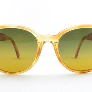 May include: Vintage-style sunglasses with a light brown frame and gradient yellow-green lenses. The round frames have small gold-colored accents on the temples. The arms are a darker brown color.