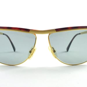 Puede incluir: Gafas de sol con montura dorada, cejas de carey y lentes azul claro. Las gafas de sol tienen un diseño retro con una parte superior curvada y un aspecto elegante y moderno. Las lentes son de color azul claro y las monturas son doradas.