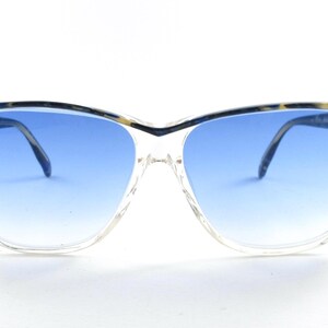 Puede incluir: Gafas de sol con montura transparente y lentes degradadas azules. La parte superior de la montura tiene un patrón decorativo con detalles florales. Las patillas son finas y oscuras. Las gafas están sobre un fondo blanco.