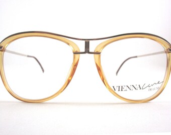May include: Vintage eyeglasses with a light brown frame and a silver-colored bridge and temples. The lenses are clear. The text "VIENNA LINE DE LUXE" is printed on the lower right lens.