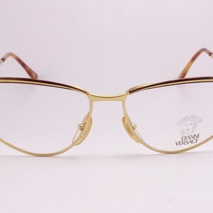 May include: Gold-tone metal eyeglasses with a brown tortoiseshell accent on the top of the frame. The glasses have a cat-eye shape and the brand name "GIANNI VERSACE" is printed on the right lens.