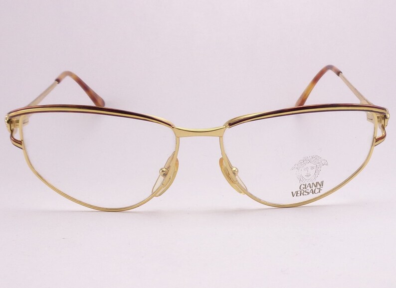 May include: Gold-tone metal eyeglasses with a brown tortoiseshell accent on the top of the frame. The glasses have a cat-eye shape and the brand name "GIANNI VERSACE" is printed on the right lens.