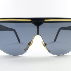 Puede incluir: Gafas de sol negras con detalles dorados y lentes azules. Las gafas tienen un diseño de escudo con una montura gruesa y un puente. Las patillas son finas y negras.