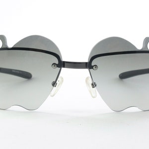 Könnte beinhalten: Ausgefallene Sonnenbrille mit schwarzem Rahmen und grauen Gläsern. Die Gläser sind wie Äpfel geformt, mit einem Stieldetail oben. Die Sonnenbrille hat ein modernes, stilvolles Design.