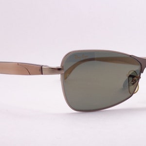 Puede incluir: Un par de gafas de sol con montura de metal marr&oacute;n y lentes verdes. Las gafas de sol tienen una etiqueta de Ray-Ban unida al brazo derecho.