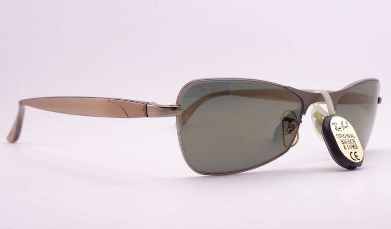 Puede incluir: Un par de gafas de sol con montura de metal marr&oacute;n y lentes verdes. Las gafas de sol tienen una etiqueta de Ray-Ban unida al brazo derecho.