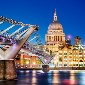 May include: A view of the Millennium Bridge in London at night, with St Paul's Cathedral in the background. The bridge is lit up with lights, and the water is reflecting the lights.