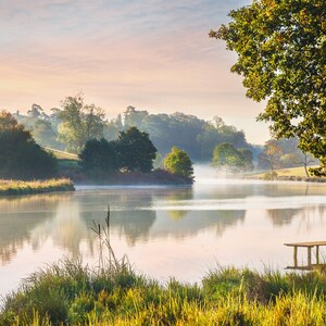 May include: A tranquil lake scene with a wooden jetty, reflecting the sky and trees. The image is bathed in soft light, with mist rising from the water. Green grass and foliage frame the foreground, creating a peaceful atmosphere.