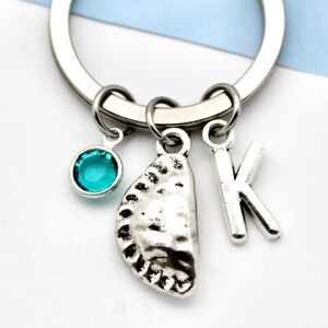 May include: Silver keyring with three charms: a teal gemstone, a textured half-circle, and the letter "K". The keyring is displayed against a white and blue backdrop.