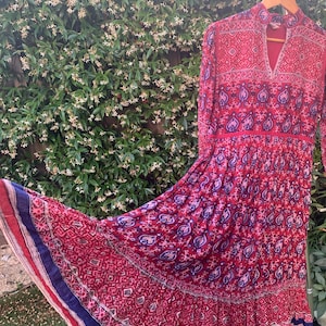 Könnte beinhalten: Ein langes, fließendes Kleid mit einem lebendigen roten und blauen Paisley-Muster. Das Kleid hat lange Ärmel, einen hohen Ausschnitt und einen gestuften Rock. Der Stoff wirkt leicht. Das Kleid hängt an einem Holzbügel.