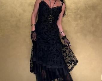 May include: A black lace dress with spaghetti straps and a layered skirt. The dress features a floral pattern and is accessorized with a cross pendant necklace, black gloves, and black boots. The background is a textured gold wall.