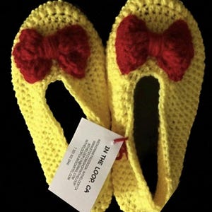 K&ouml;nnte beinhalten: Ein Paar gelbe H&auml;kelpantoffeln mit roten Schleifen. Die Pantoffeln sind handgefertigt und haben ein Etikett mit der Aufschrift "IN THE LOOP. CA".