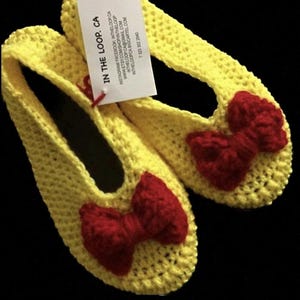 K&ouml;nnte beinhalten: Ein Paar gelbe H&auml;kelpantoffeln mit roten Schleifen. Die Pantoffeln sind handgefertigt und bieten einen bequemen Sitz.