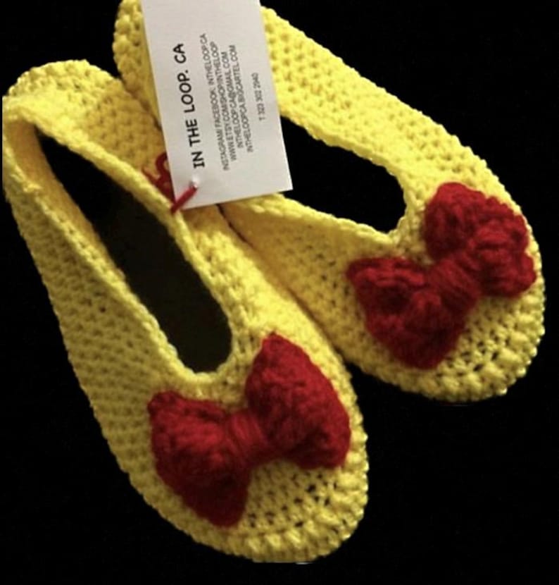 K&ouml;nnte beinhalten: Ein Paar gelbe H&auml;kelpantoffeln mit roten Schleifen. Die Pantoffeln sind handgefertigt und bieten einen bequemen Sitz.