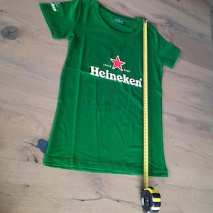 May include: Green t-shirt with the Heineken logo, featuring a red star and white text. A yellow measuring tape is placed next to the shirt. The shirt is approximately 36 inches long.