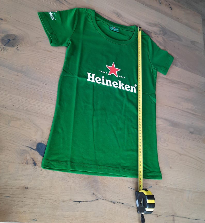 May include: Green t-shirt with the Heineken logo, featuring a red star and white text. A yellow measuring tape is placed next to the shirt. The shirt is approximately 36 inches long.