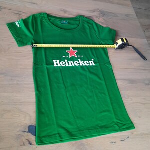 May include: Green t-shirt with the Heineken logo, featuring a red star and white text. The shirt is measured with a tape measure, showing a width of approximately 18 inches. The shirt has short sleeves and a crew neck.