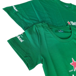 May include: Two green t-shirts with the Heineken logo, featuring a red star and white text. The shirts are made of a soft material and have the word "Heineken" printed on the front. The sleeve has the word "Eken" printed on it.