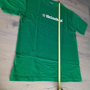 May include: A green t-shirt with the Heineken logo in white and a red star. The shirt is measured with a tape measure, showing a length of approximately 30 inches.