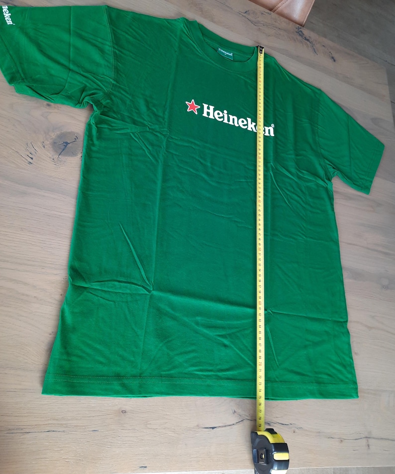 May include: A green t-shirt with the Heineken logo in white and a red star. The shirt is measured with a tape measure, showing a length of approximately 30 inches.
