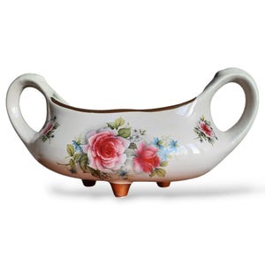 Puede incluir: Plato de servir de cerámica blanca con tres patas y dos asas. El plato tiene un diseño floral con rosas rosas y rojas y hojas verdes.