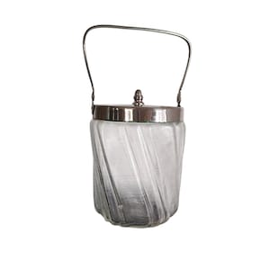 Op de afbeelding: Een heldere glazen pot met een zilverkleurige deksel en handvat. De pot heeft een textuur met een wervelend ontwerp. Het handvat is een eenvoudige, gebogen metalen staaf. De deksel heeft een kleine, decoratieve knop.