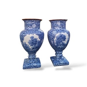 May include: A pair of blue and white ceramic urns featuring a detailed landscape scene with trees on the central panel, surrounded by intricate geometric patterns on the neck, base, and sides, with a wide flared rim and a square pedestal base.