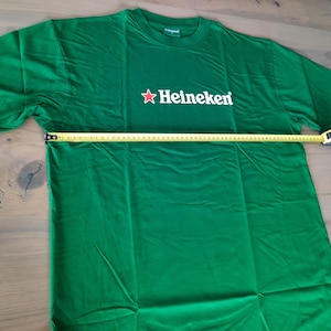 May include: Green t-shirt with the Heineken logo in white and a red star. The shirt is being measured with a tape measure. The shirt is approximately 30 inches wide.