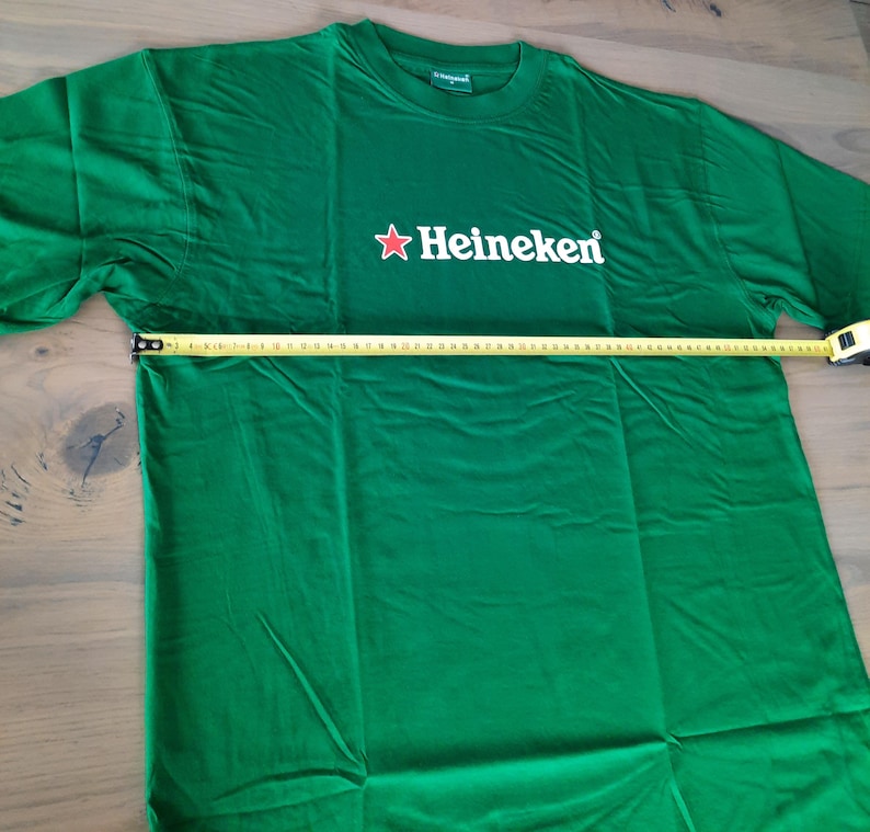 May include: Green t-shirt with the Heineken logo in white and a red star. The shirt is being measured with a tape measure. The shirt is approximately 30 inches wide.